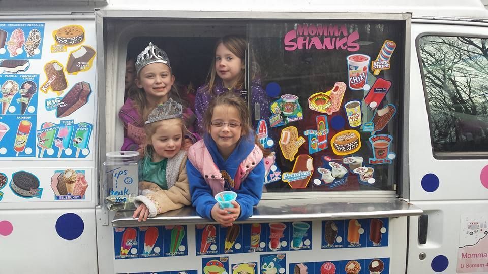 Four children smile through the serving window of a white ice cream truck decorated with pictures of treats.