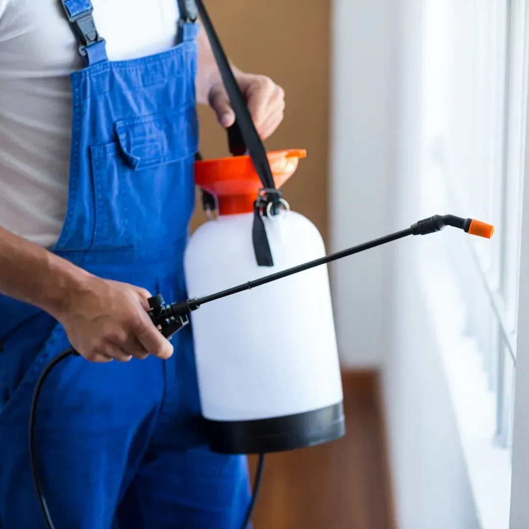 Person in blue overalls holding a garden sprayer with a black wand indoors
