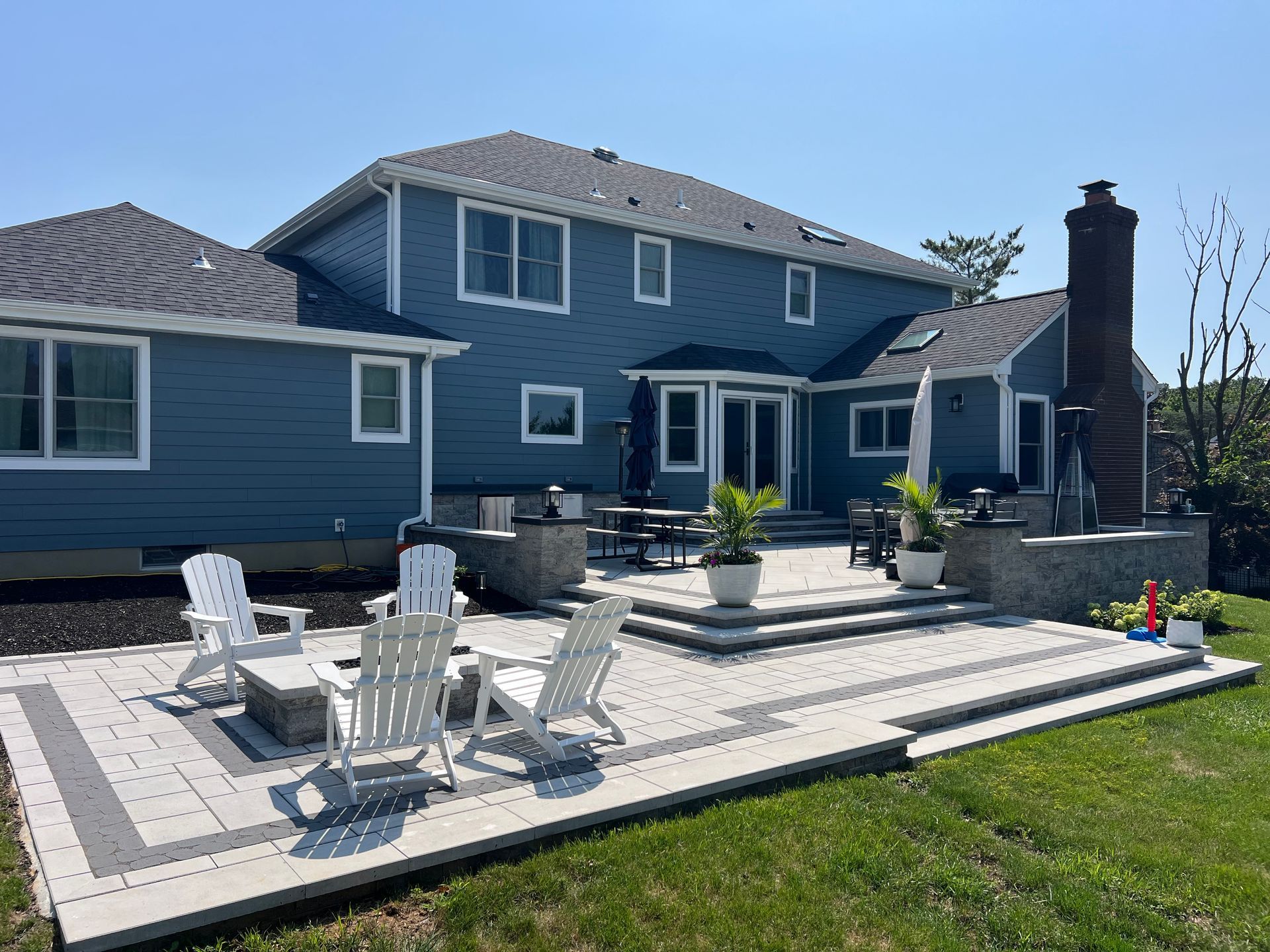 Blue house with stone patio, white chairs, and potted plants in a sunny backyard