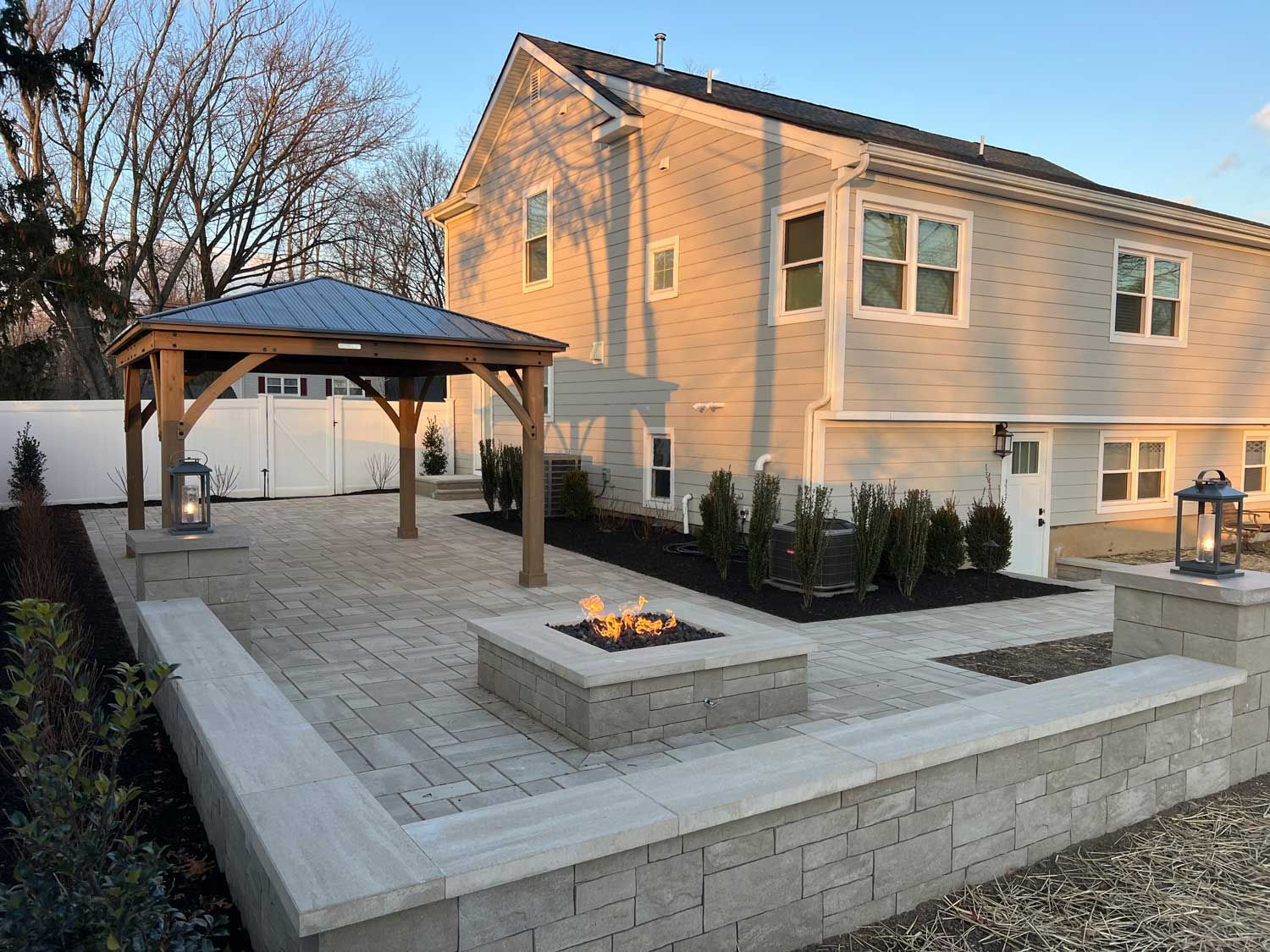 Backyard patio with pergola, fire pit, and raised stone seating wall beside a two-story house