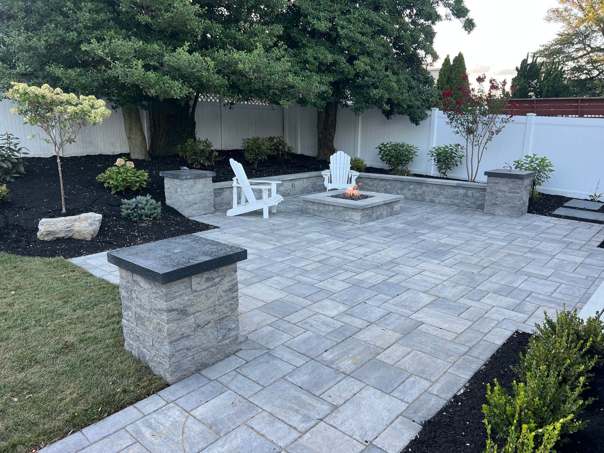 Backyard patio with fire pit, white chairs, gray pavers, black mulch, and greenery.