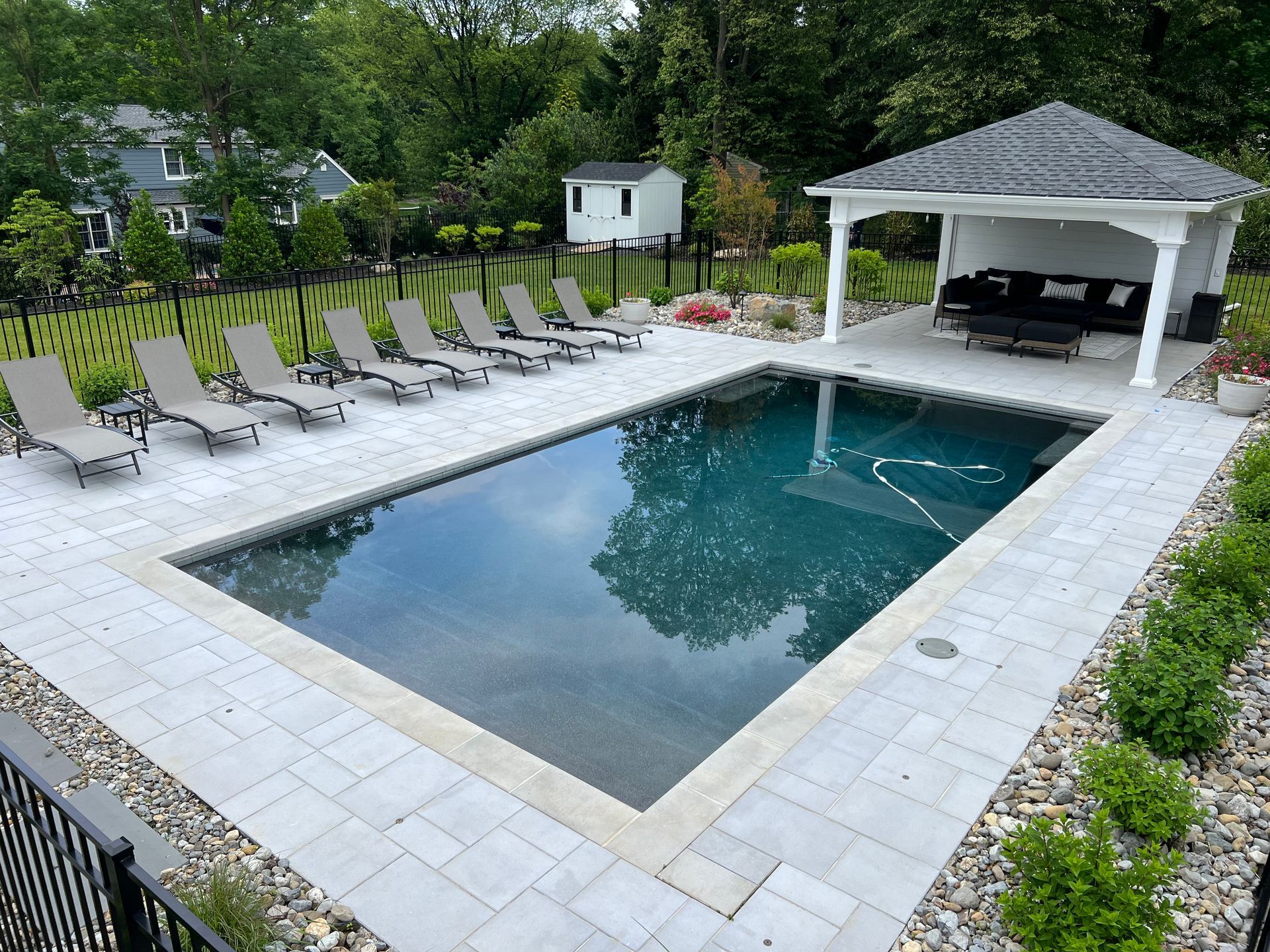 A rectangular pool with a gazebo, lounge chairs, and landscaping. Green trees and a house are in the background.
