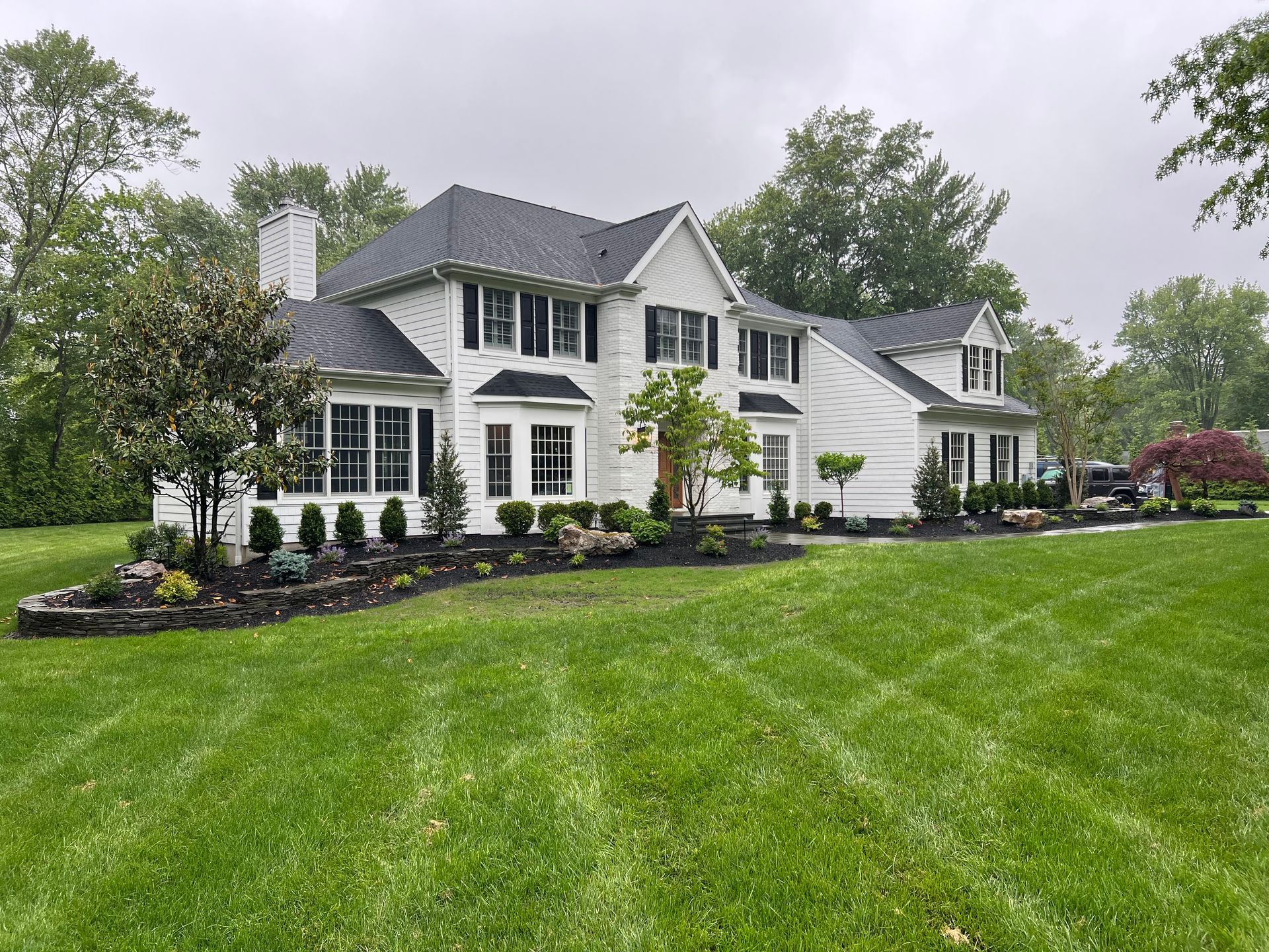 White two-story house with black shutters on a grassy lawn, surrounded by trees.