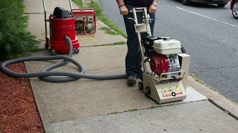 Person using a concrete grinder on a sidewalk, vacuum connected. Red engine, generator, parked car.