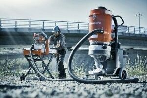 Construction worker using a saw, connected to an orange vacuum, near a bridge.