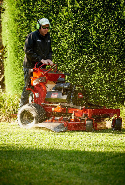 Man mowing a lawn with a red ride-on mower, wearing ear protection, in a yard with a green hedge.