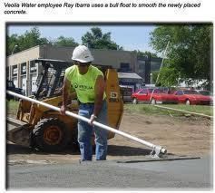 Veolia Water employee Ray Ibarra using a bull float to smooth fresh concrete near a building.