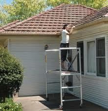 Person on a scaffold cleaning gutters on a white house with a brown roof.