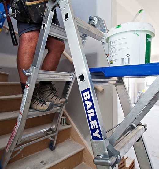 A person stands on a Bailey adjustable ladder on stairs, with a paint bucket and tray nearby.