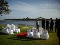 Wedding setup outdoors with chairs, red carpet, and water background; guests nearby.