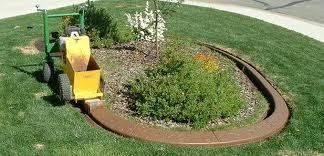 A yellow machine is creating a brown concrete border around a flower bed in green grass.