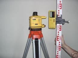 Yellow surveying equipment on a tripod, with a leveling rod held by a hand, indoors.