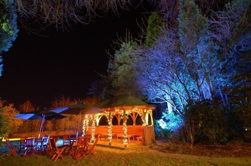 Nighttime gazebo illuminated by lights, with tables and chairs on a lawn, surrounded by trees with blue lighting.