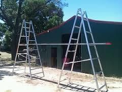 Two aluminum ladders supporting a horizontal beam in front of a green building.