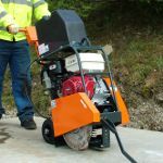 Person operating an orange concrete saw; outdoors, near a sidewalk.
