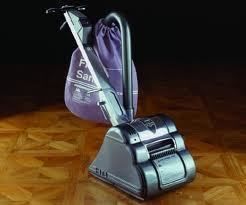Upright vacuum cleaner with purple bag, on a wood floor, against a dark background.