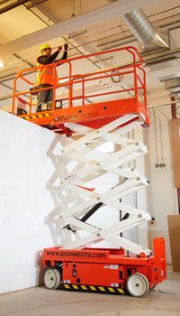 Worker on orange scissor lift, reaching towards ceiling in a warehouse.