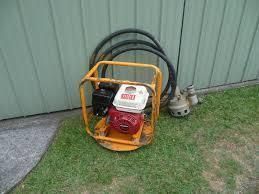 Yellow plate compactor, black hoses, and a small pump on green grass.