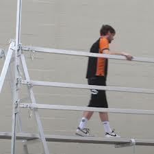 Man walking on metal scaffolding, wearing black and orange shirt, black shorts, and sneakers.