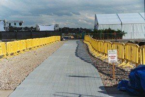 Pathway lined with yellow barriers, leading to a white tent on gravel and concrete. Overcast sky.