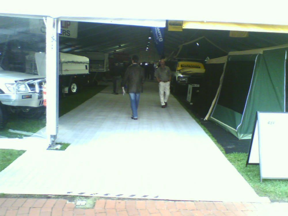 People walking through an outdoor trade show, with tents, vehicles, and displays visible.