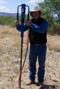 Man in cowboy hat operating a blue post driver, outdoors in a field.