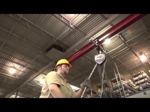 Man in hardhat operating an overhead hoist with a chain, inside a factory.