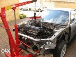 Red engine hoist lifting a car engine out of a silver car in an outdoor setting.