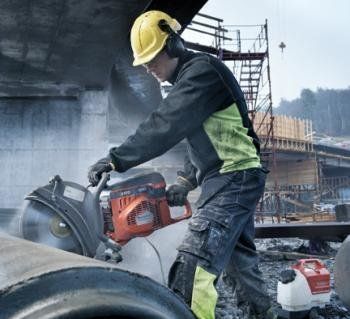 Construction worker wearing safety gear, cutting through a pipe with a power saw at a construction site.