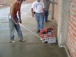 Three construction workers smoothing concrete floor with a power tamper in a building with brick walls.