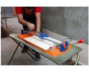 Man using a tile cutter on a light-colored tile, outdoors, near a wall.