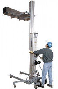 Person operating a material lift, raising a rectangular item. He wears a hard hat and jeans in a neutral setting.