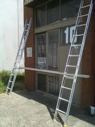 Two aluminum ladders lean against a building with a window. A white board rests on the sill.