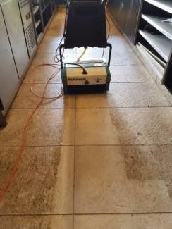 Floor cleaner scrubbing a dirty tile floor between kitchen cabinets.