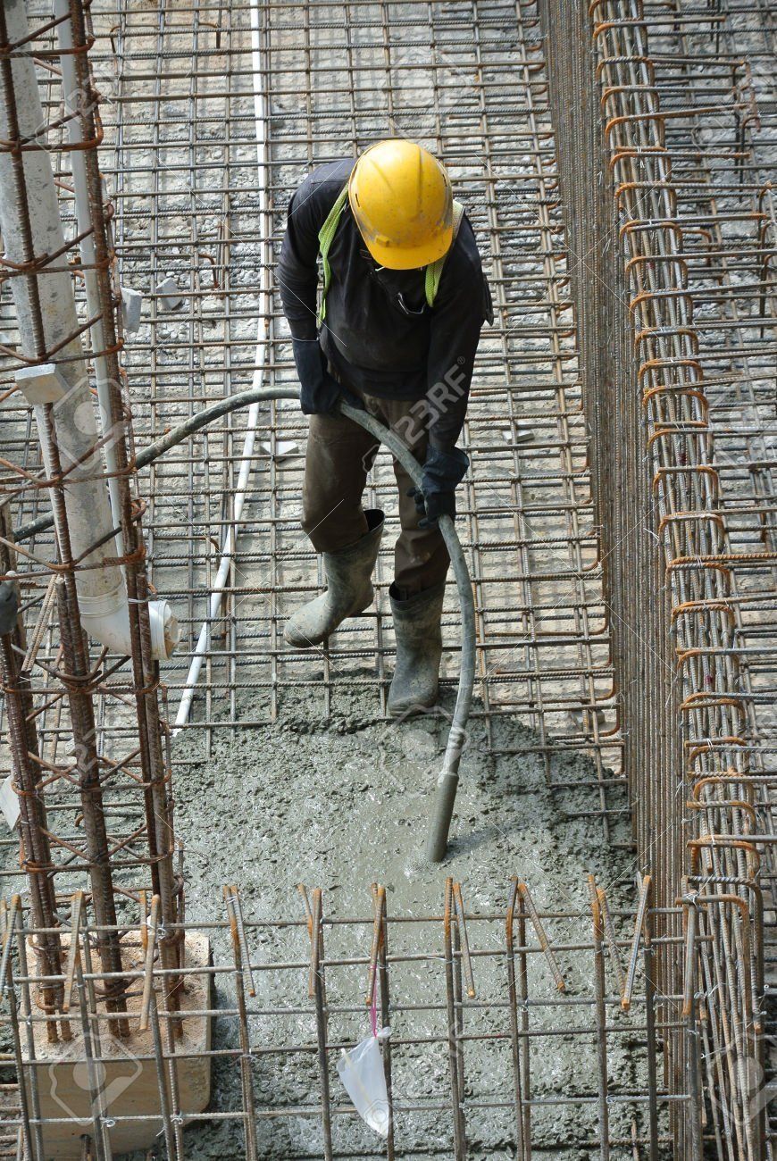 Construction worker using a vibrator on wet concrete within a steel rebar frame. He's wearing a yellow helmet and boots.
