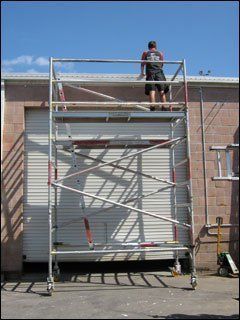 Person on a tall rolling scaffold in front of a building with a closed garage door.