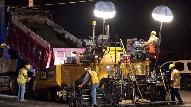 Road construction at night: A dump truck unloading asphalt into a paver, workers in vests and hard hats.