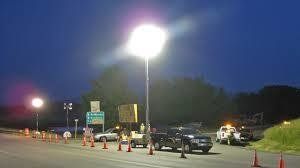 Night scene with bright lights illuminating a road, traffic cones, vehicles, and highway signs.