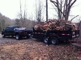 Black truck towing a trailer loaded with firewood on a dirt road in a wooded area.