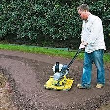 Man compacts gravel path with a yellow plate compactor in a grassy outdoor area.