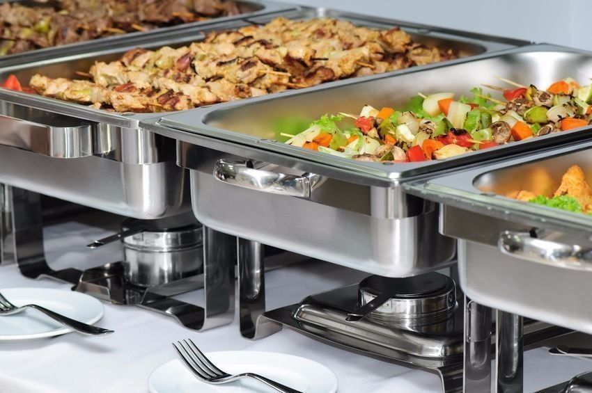Buffet table with several chafing dishes filled with various cooked food, on a white tablecloth.