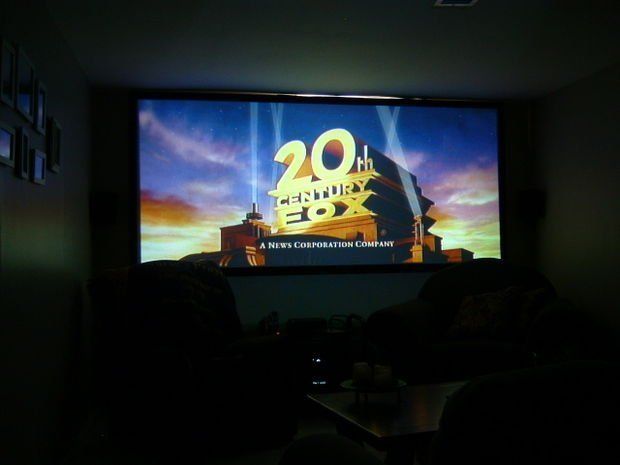 20th Century Fox logo on a large screen in a home theater; silhouetted figures in the foreground.