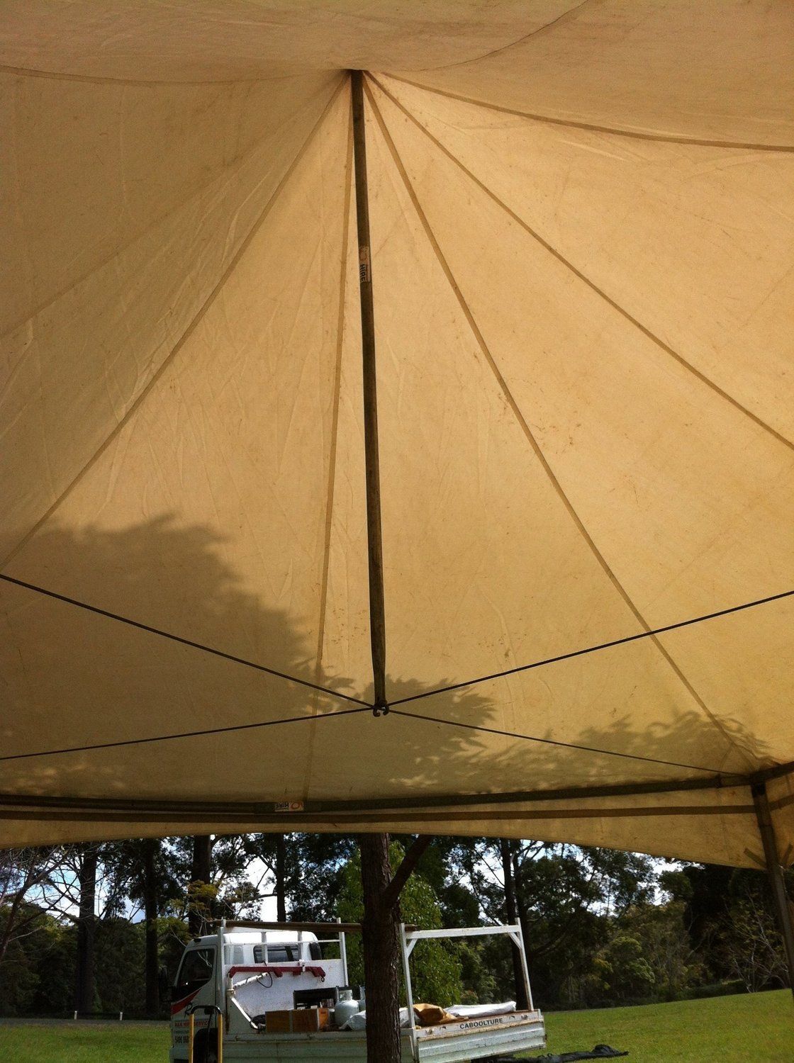 View from under a tan tent, looking up at the central support pole and converging fabric. A vehicle is visible below.