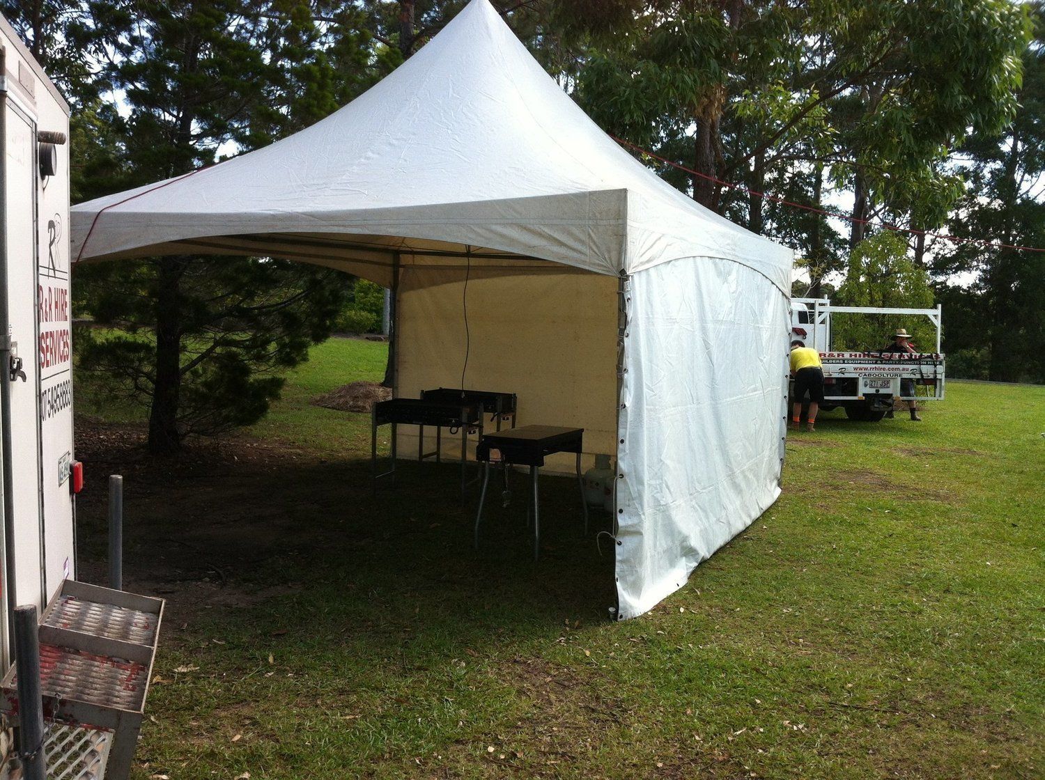 White tent with grills on green grass, parked truck, and people setting up outdoors.