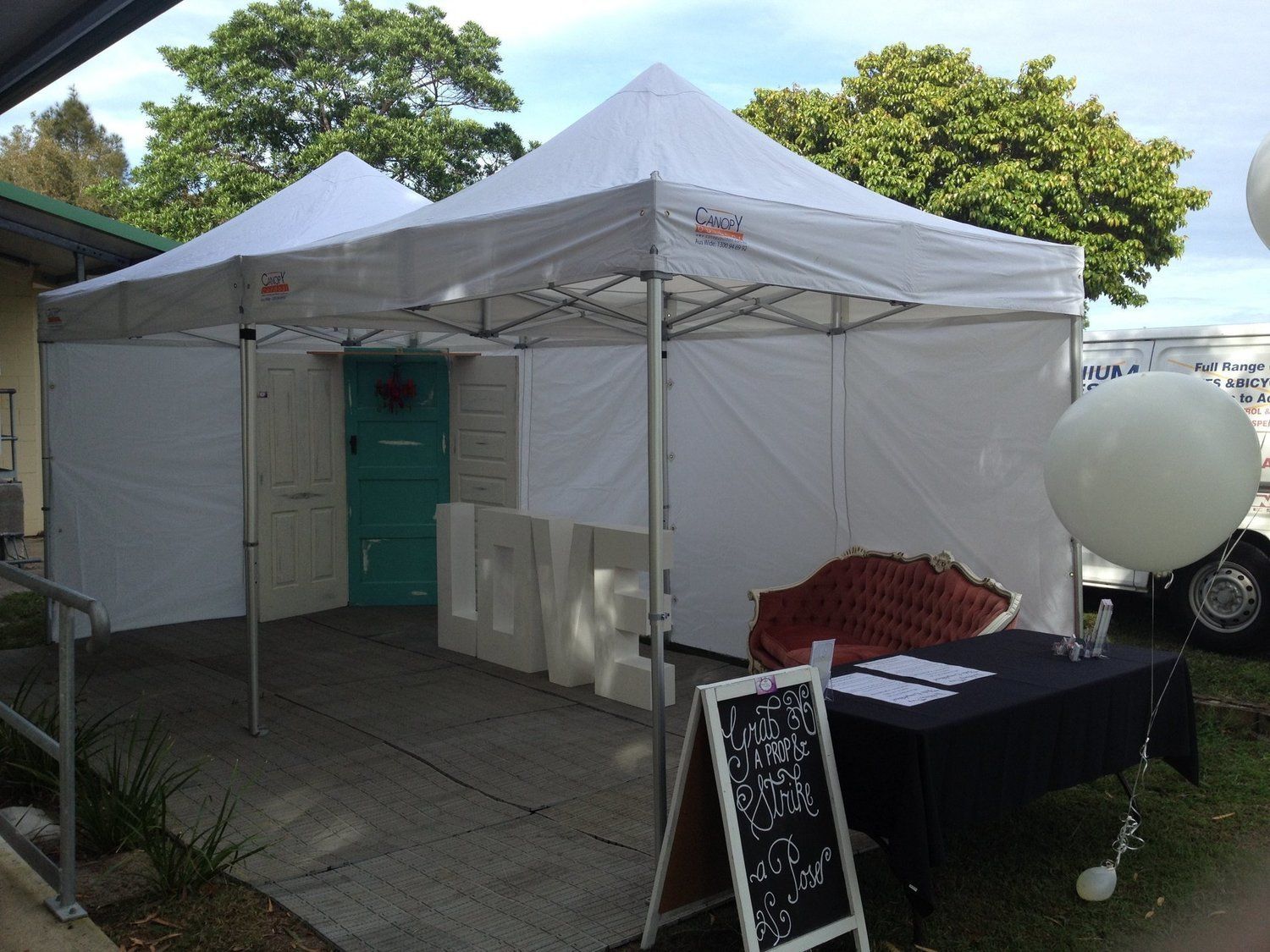 White pop-up tents enclose a teal door, with a table, red loveseat, and chalkboard sign.