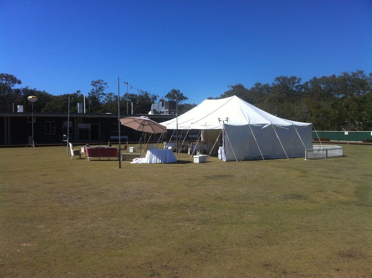 White tent set up on a green lawn under a clear blue sky, with a building and trees in the background.