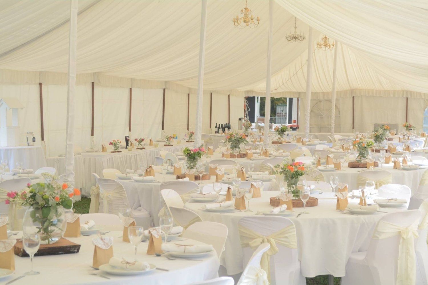 White tent interior set for a wedding reception; round tables with floral centerpieces, chairs draped in white.