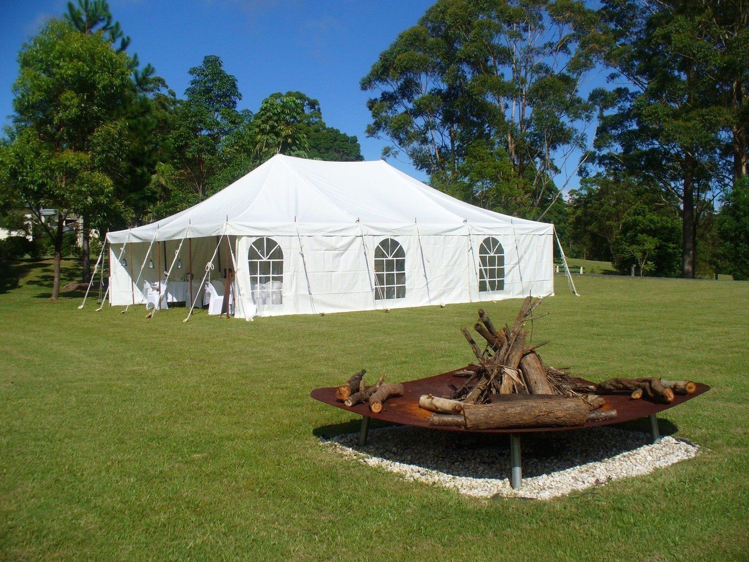 White tent set up on green grass with a fire pit and trees in the background.