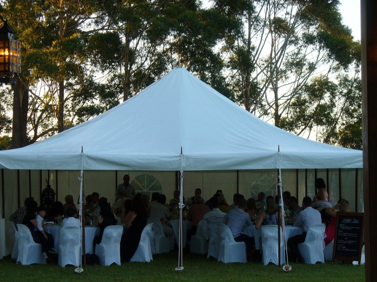 White tent with tables and people, set outdoors with trees in the background.