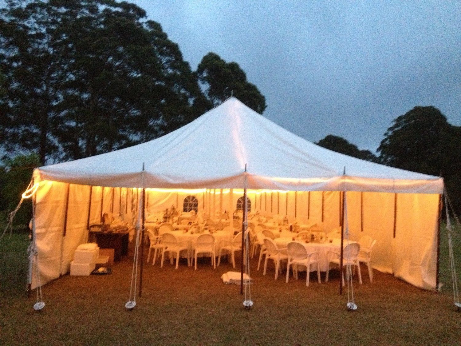 White tent set up on grass, lit from within, with tables and chairs arranged inside; outdoor setting at dusk.